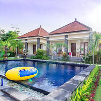 View of a serene outdoor pool with a yellow inflatable raft, surrounded by tropical landscaping and a villa-style house with red-tiled roofs in the background.