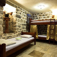 Room with stone walls featuring a single bed and a bunk bed with a ladder. There's a ceiling light and two cushions on the single bed.