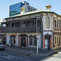 A historic two-story building with a balcony and ironwork railings, situated on a street corner. Modern high-rise buildings are visible in the background.