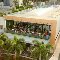 Aerial view of a two-story building with a crowded outdoor seating area on the upper floor, surrounded by palm trees.