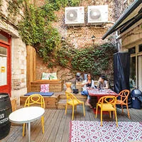 Outdoor cafe with three people sitting at a table. Colorful chairs and rugs enhance the cozy setting, surrounded by brick walls with greenery and air conditioning units above.