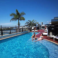 A couple sits by a rooftop pool with clear blue water, surrounded by lounge chairs and palm trees. A cityscape is visible in the background under a clear blue sky.