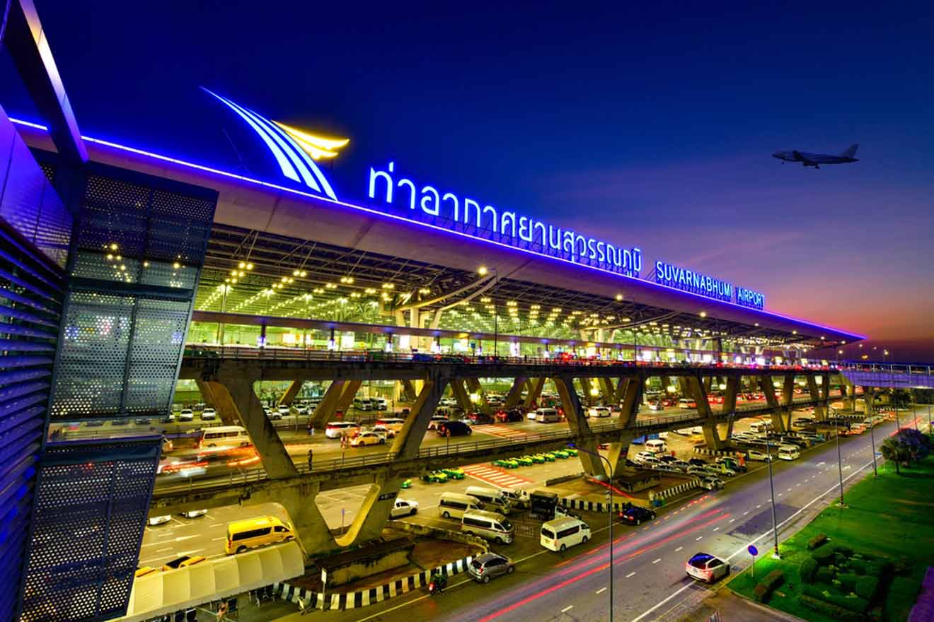 Exterior view of Suvarnabhumi Airport at dusk, with bright signage and an airplane flying overhead. Multiple cars are visible in the busy foreground.