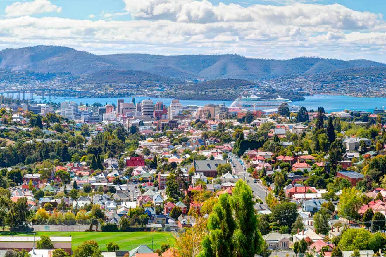 Aerial view of a cityscape with colorful rooftops, surrounded by green hills and a distant body of water under a partly cloudy sky.