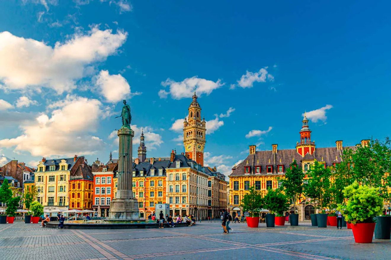 A vibrant city square with colorful historic buildings, a central monument, and a clock tower under a blue sky with clouds. People and large red planters are scattered around the square.