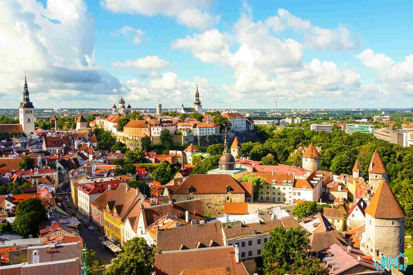 View of a historic cityscape with red-roofed buildings and a tall church spire surrounded by trees under a clear blue sky.