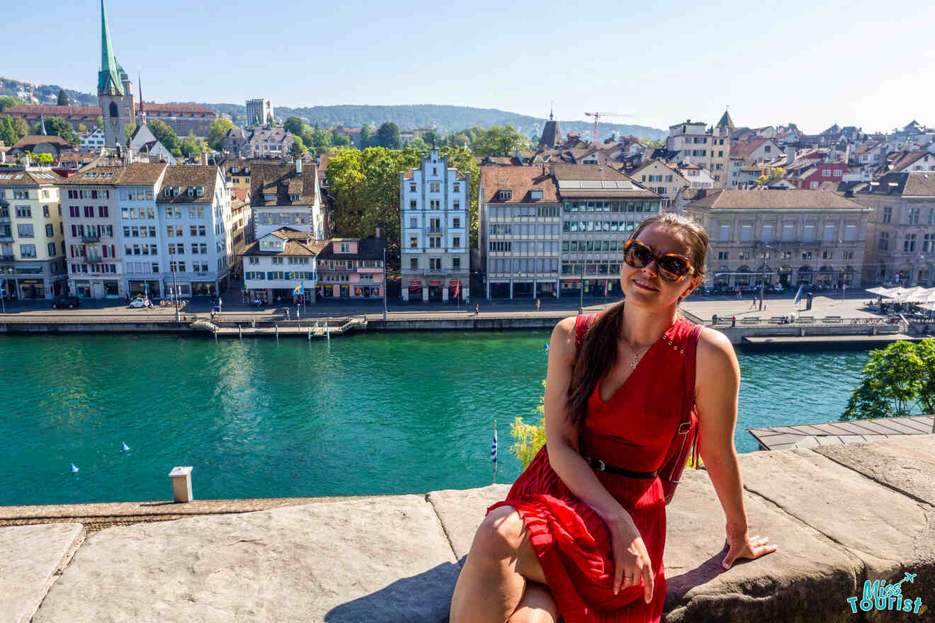 Yulia, the founder of this website, in a red dress and sunglasses sits by a river with a panoramic view of a European cityscape in the background.