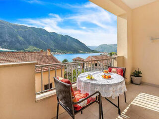 A balcony with a table set for two, overlooking a rooftop and a scenic view of mountains and a lake under a clear blue sky.