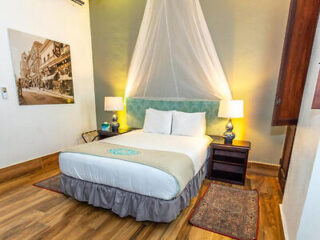 A cozy bedroom with a double bed, mosquito net, two bedside lamps, and wooden flooring. A framed photograph hangs on the wall.