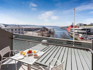 Balcony view of a harbor with docked ships, buildings, and distant hills. A small table holds a breakfast setup with two chairs.