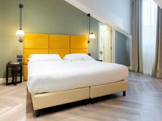 A modern bedroom with a large bed featuring a yellow headboard, white bedding, two wall-mounted lamps, and a window with beige curtains.