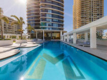 Outdoor pool in front of a modern high-rise building, with palm trees and lounge chairs along the sides.