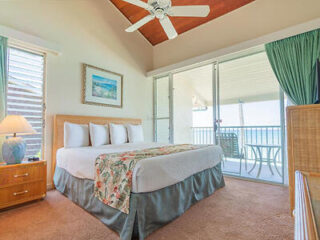 A bedroom with a large bed, three pillows, and a floral throw. There is a ceiling fan, a wooden dresser, a table lamp, and sliding glass doors leading to a balcony with a table and chair.