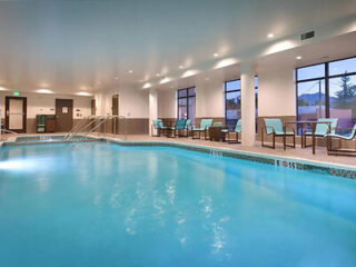 Indoor swimming pool with clear blue water, surrounded by lounge chairs and large windows. Ceiling lights illuminate the area.