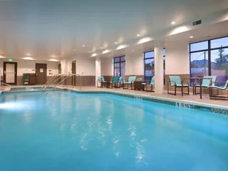 Indoor swimming pool with clear blue water, surrounded by lounge chairs and large windows. Ceiling lights illuminate the area.