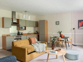 Modern living room with mustard sofa, cozy throw, and small dining area. The open kitchen features light wood cabinets, stainless steel appliances, and a wall-mounted microwave.