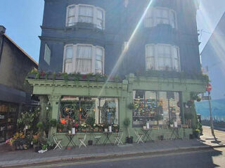 Street view of a green corner building with a floral shop and several tables and chairs outside, under bright sunlight.