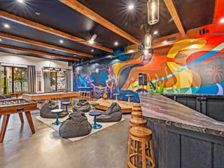 Colorful lounge area with a foosball table, bean bag seating, stools at a bar counter, and a vibrant wall mural.