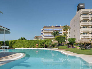 Outdoor hotel pool with surrounding greenery and sun loungers, adjacent to a multi-story building under clear blue sky.