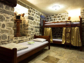 A rustic room with stone walls, containing a single bed and a bunk bed with a ladder. Pillows and folded towels are on the beds. The floor is tiled, and there is soft lighting.