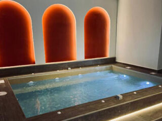 A rectangular indoor jacuzzi features bubbling water, surrounded by dark tiles. Behind it, three large, arched niches glow with orange lighting.