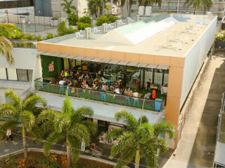 Aerial view of a building with a rooftop terrace where people are seated at tables. Palm trees border the structure, and a walkway runs alongside the building.