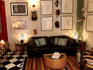 A cozy living room with a black sofa, patterned pillows, and a wooden coffee table. The walls feature framed art and masks. The floor has a colorful striped rug and checkerboard tiles.