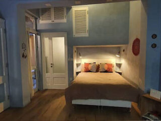 Cozy bedroom with a double bed, bedside lamps, and decorative pillows. A door to the left leads to a different room. Walls are painted in light blue and white.