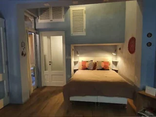 Cozy bedroom with a double bed, bedside lamps, and decorative pillows. A door to the left leads to a different room. Walls are painted in light blue and white.