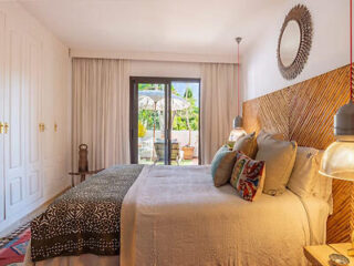 A cozy bedroom with a double bed, decorative pillows, and a patterned blanket. There's a large mirror on the wooden headboard and a sliding door leading to a sunlit patio with greenery.