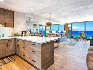 Modern open-concept kitchen and living room with wooden flooring, sleek cabinets, and a large window showcasing an ocean view. Cozy seating area near the window.