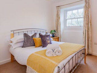 A cozy bedroom with a double bed, yellow throw, and gray and yellow pillows. A window with curtains is in the background, and a potted plant sits on a wooden nightstand. Towels are on the bed.