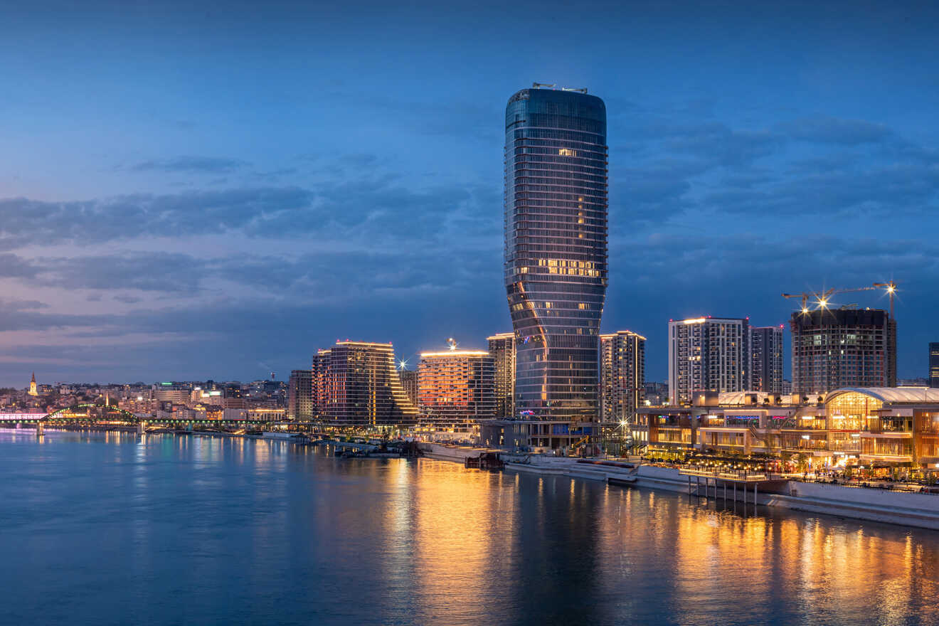 A modern city skyline at dusk is reflected in a calm body of water. The scene features a distinctive, curved high-rise building among other illuminated structures.