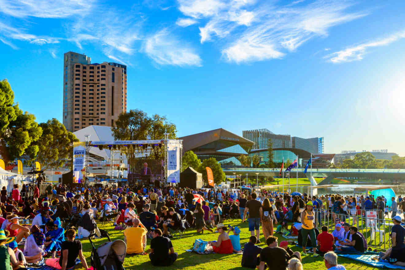 A large crowd gathers on a grassy area for an outdoor event. A stage is set up with people seated and standing. Buildings and a river are visible in the background under a clear blue sky.