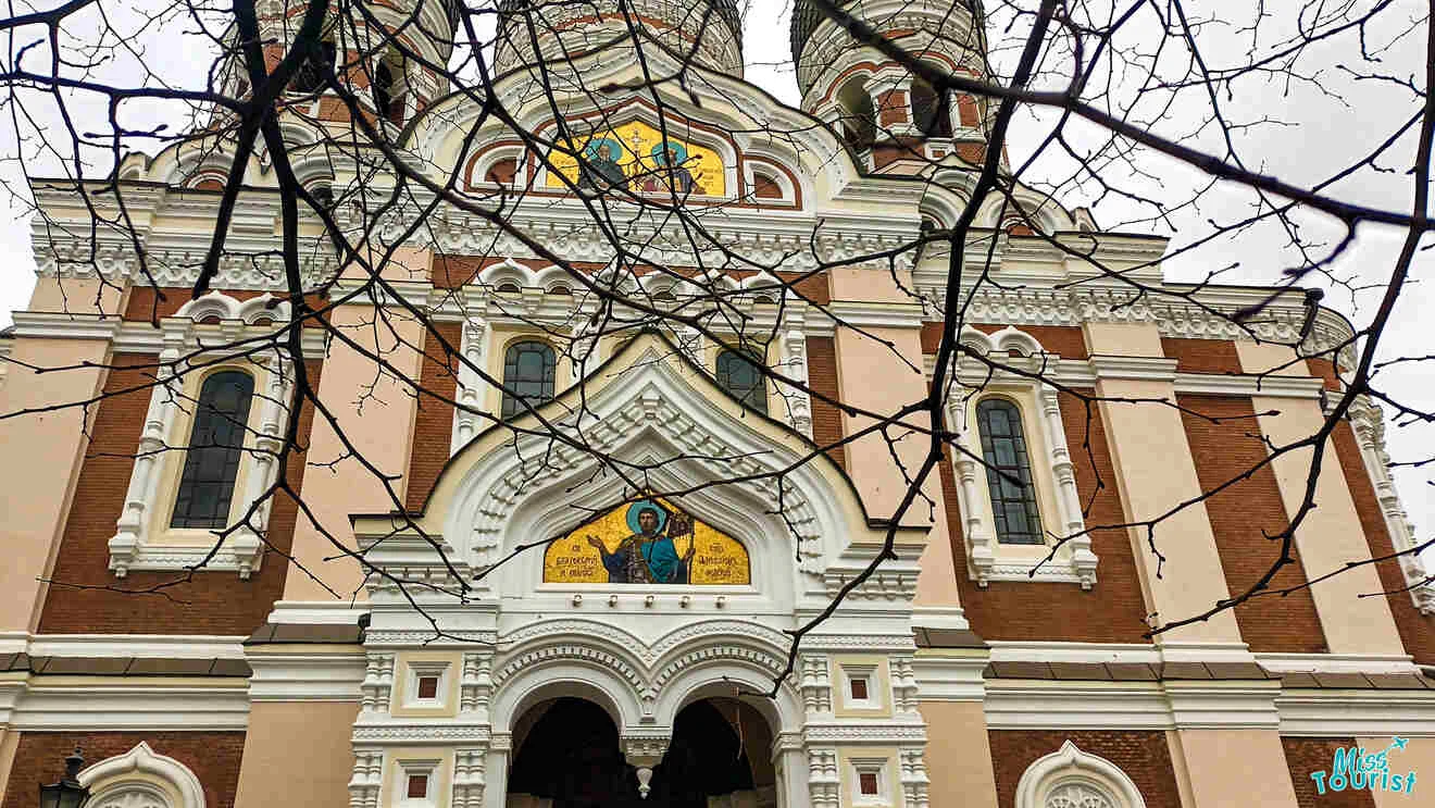 Facade of a historic building with ornate architecture, featuring religious artwork above the entrance and bare tree branches in the foreground.