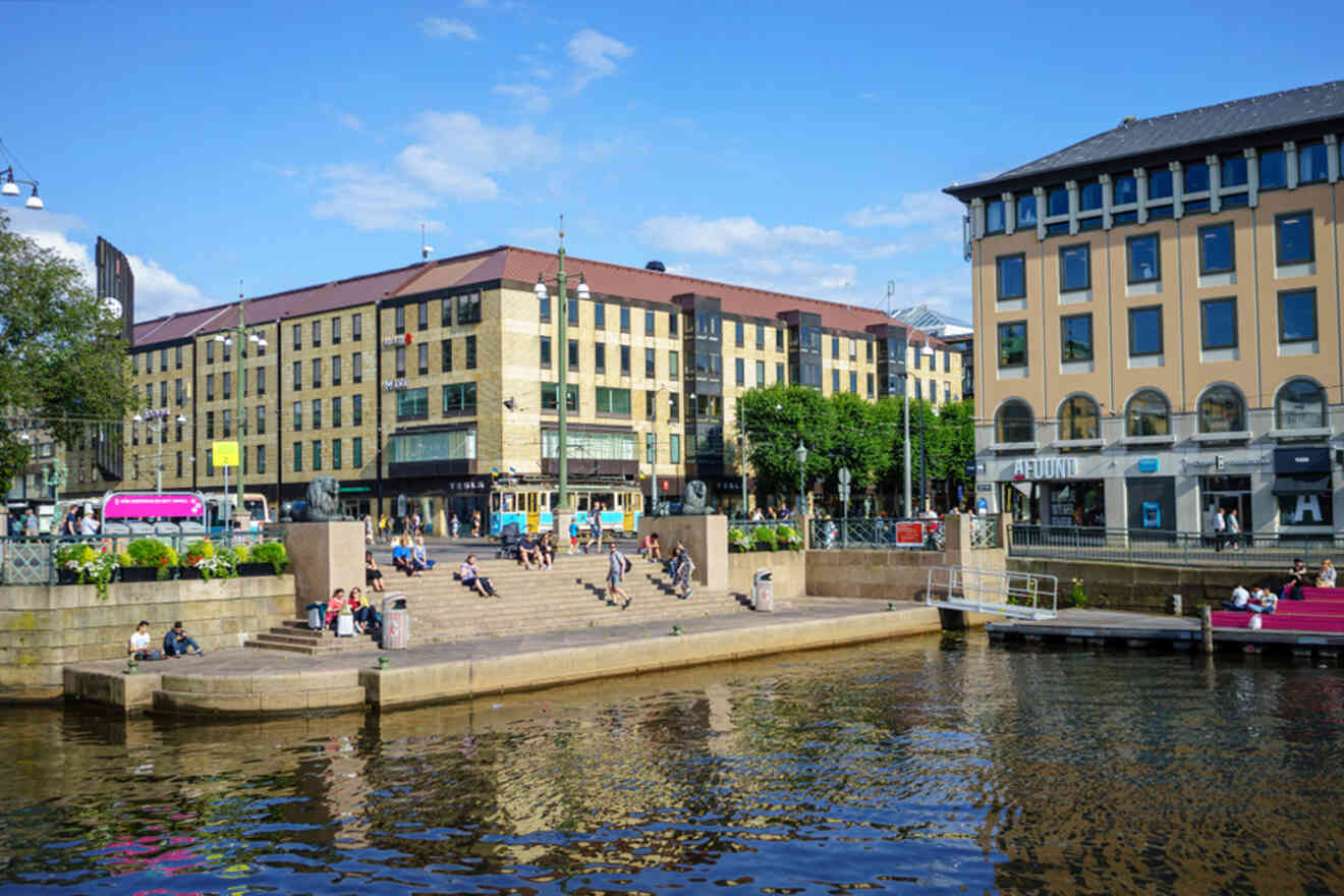 People sit on steps by a waterfront in an urban area with modern buildings under a clear blue sky.