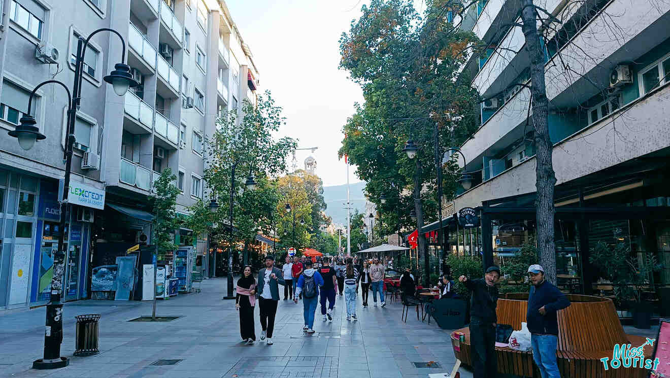 Busy pedestrian street lined with shops and cafes, people walking and sitting outside, trees on either side, and a clock tower in the background.