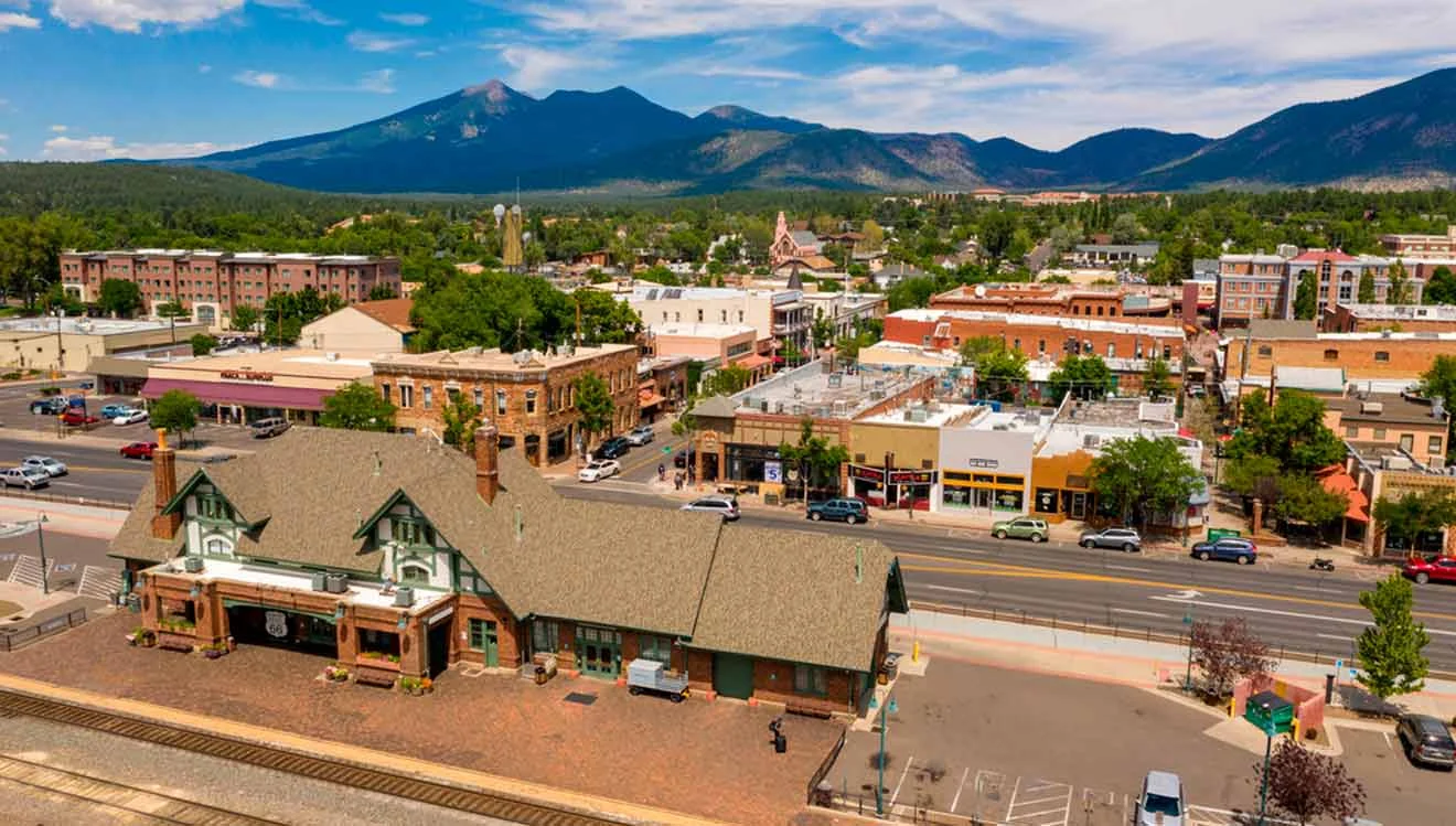 Aerial view of a small town with a railway station in the foreground, buildings, and tree-lined streets, set against a backdrop of mountains under a partly cloudy sky.