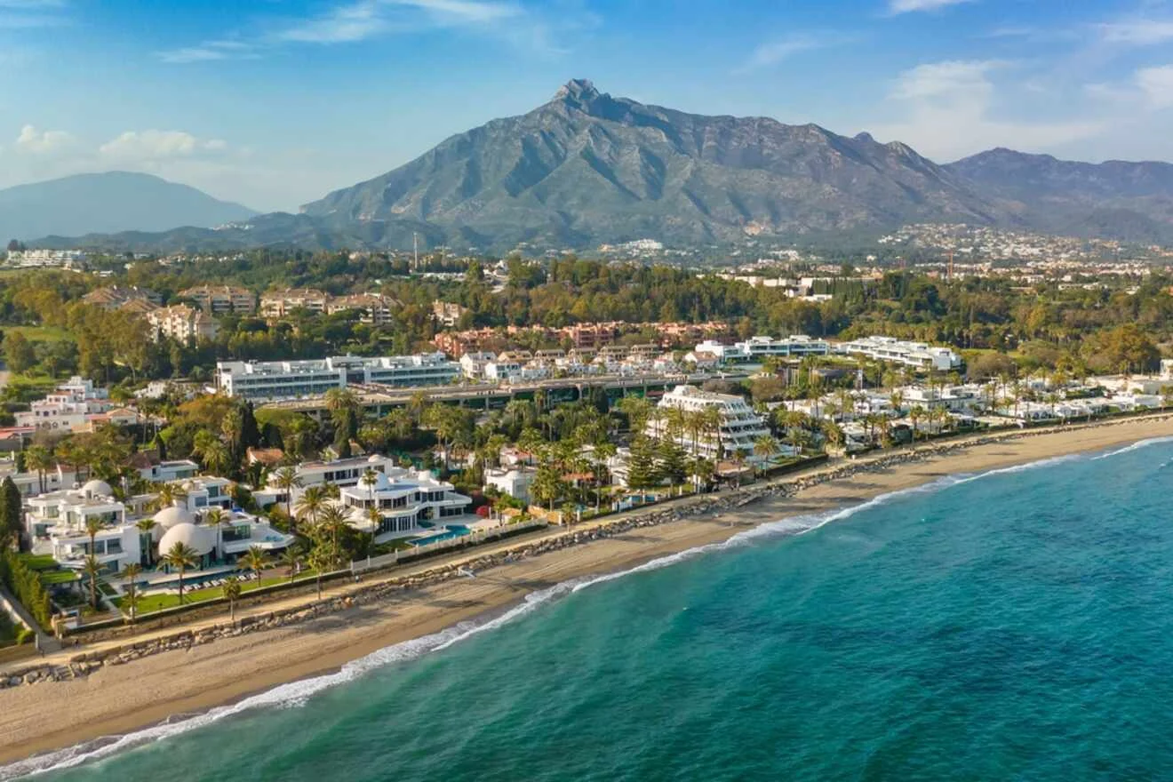 Aerial view of a coastal town with white buildings, a sandy beach, and a mountainous backdrop under a clear blue sky.