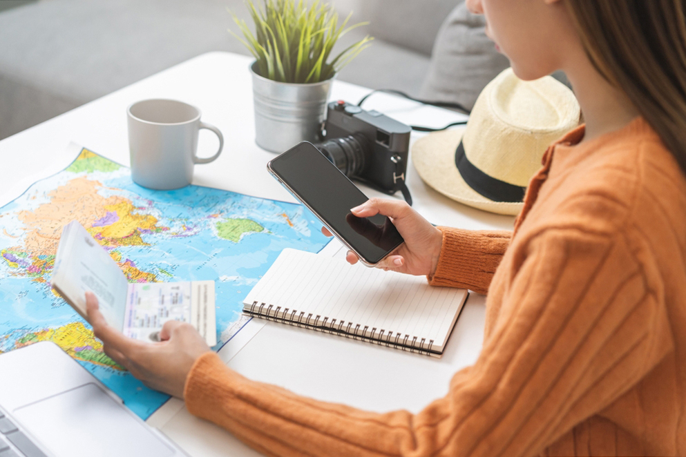 A person in an orange sweater holds a smartphone and a passport over a desk with a map, notebook, camera, mug, and plant.