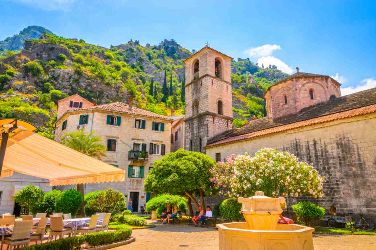 Scenic view of a historic European town square with a fountain, surrounded by stone buildings, lush greenery, and a mountainous backdrop under a clear blue sky.