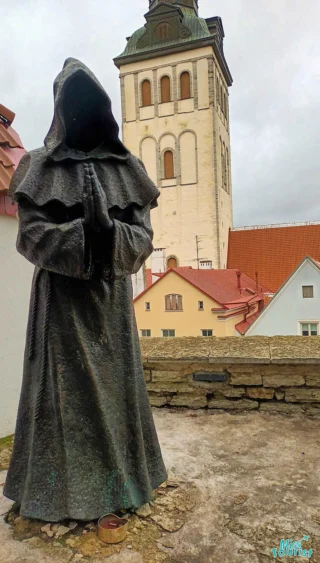 Bronze statue of a hooded figure with hands in prayer, set against a backdrop of a stone tower and rooftops under a cloudy sky.