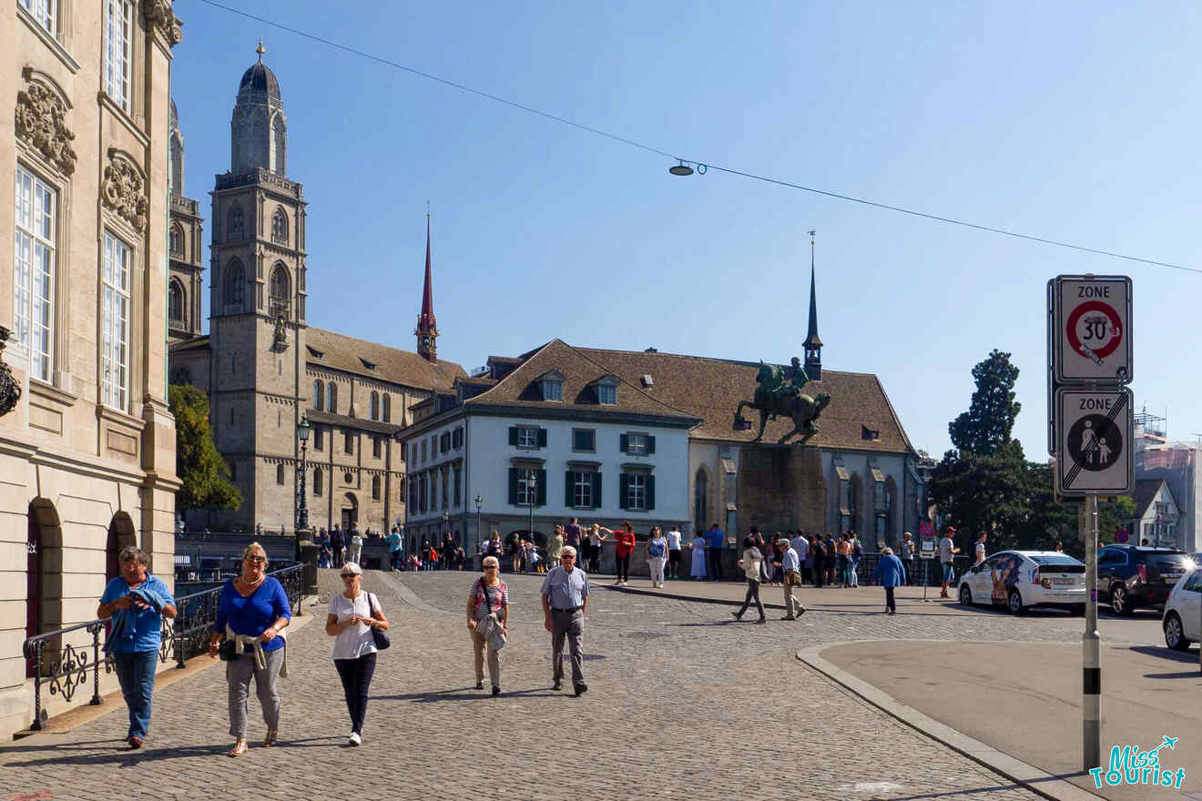 People walk on a cobblestone street near historic buildings and a church tower in a European city. Traffic signs are visible on the right.