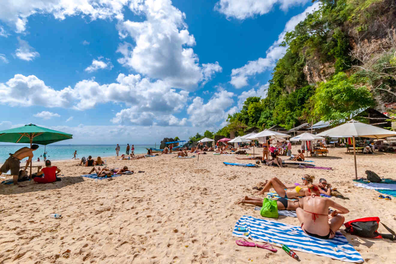 People relax on a sandy beach under umbrellas, with green cliffs in the background and a partly cloudy sky.