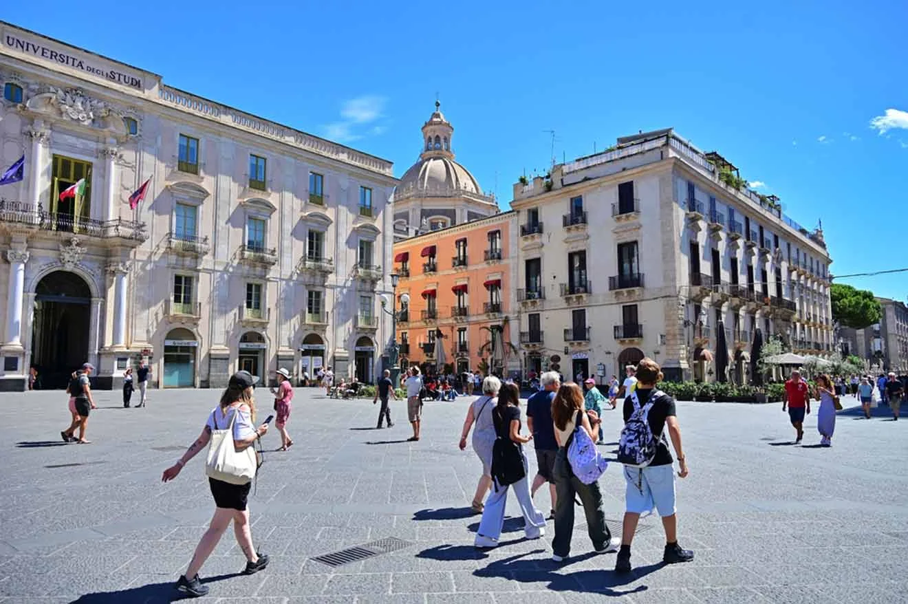 People walk in a sunny plaza with historic buildings and a domed church in the background.