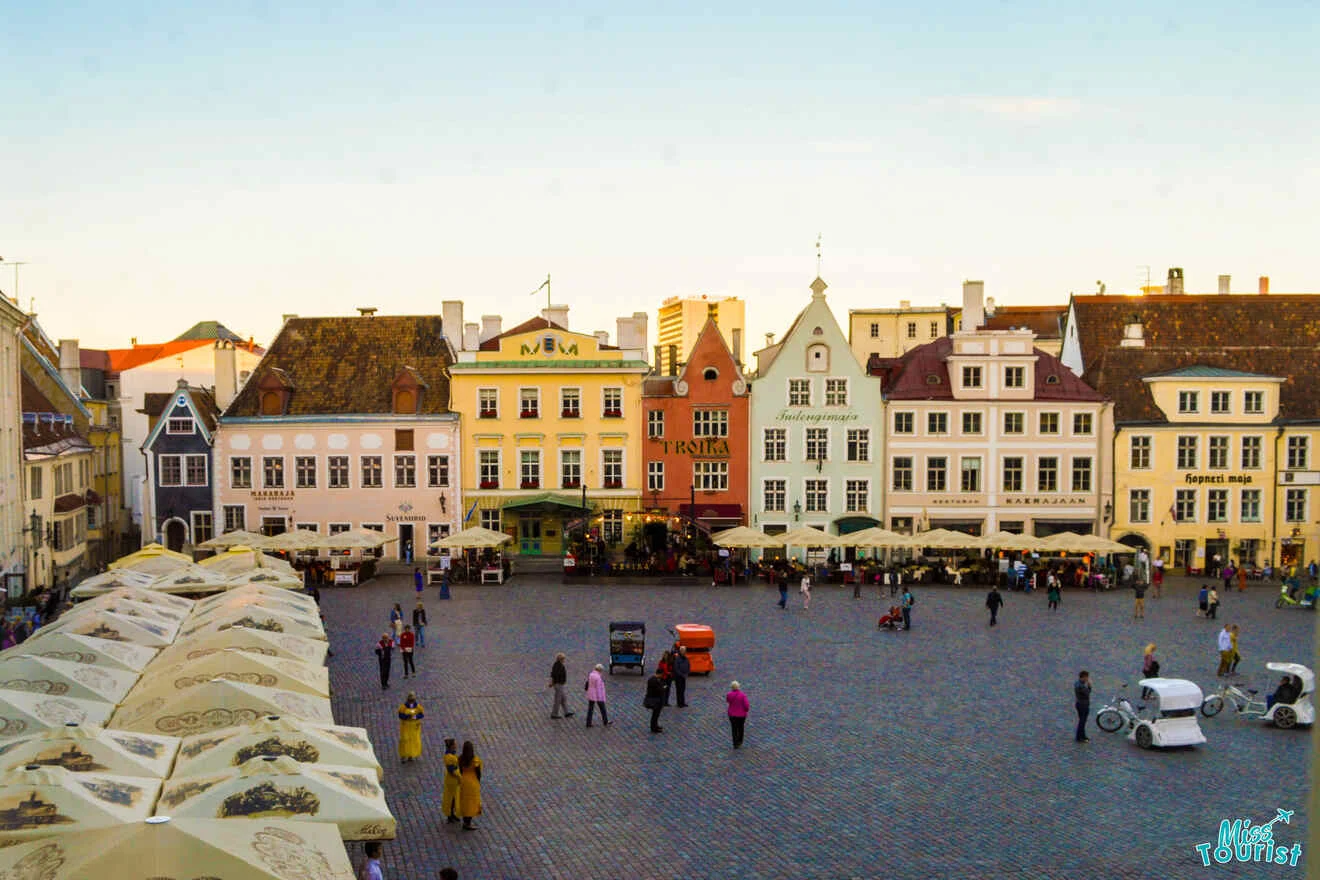 Aerial view of a European square surrounded by colorful historic buildings. People are walking and sitting, and there are canopies and small vehicles parked on the cobblestone ground.