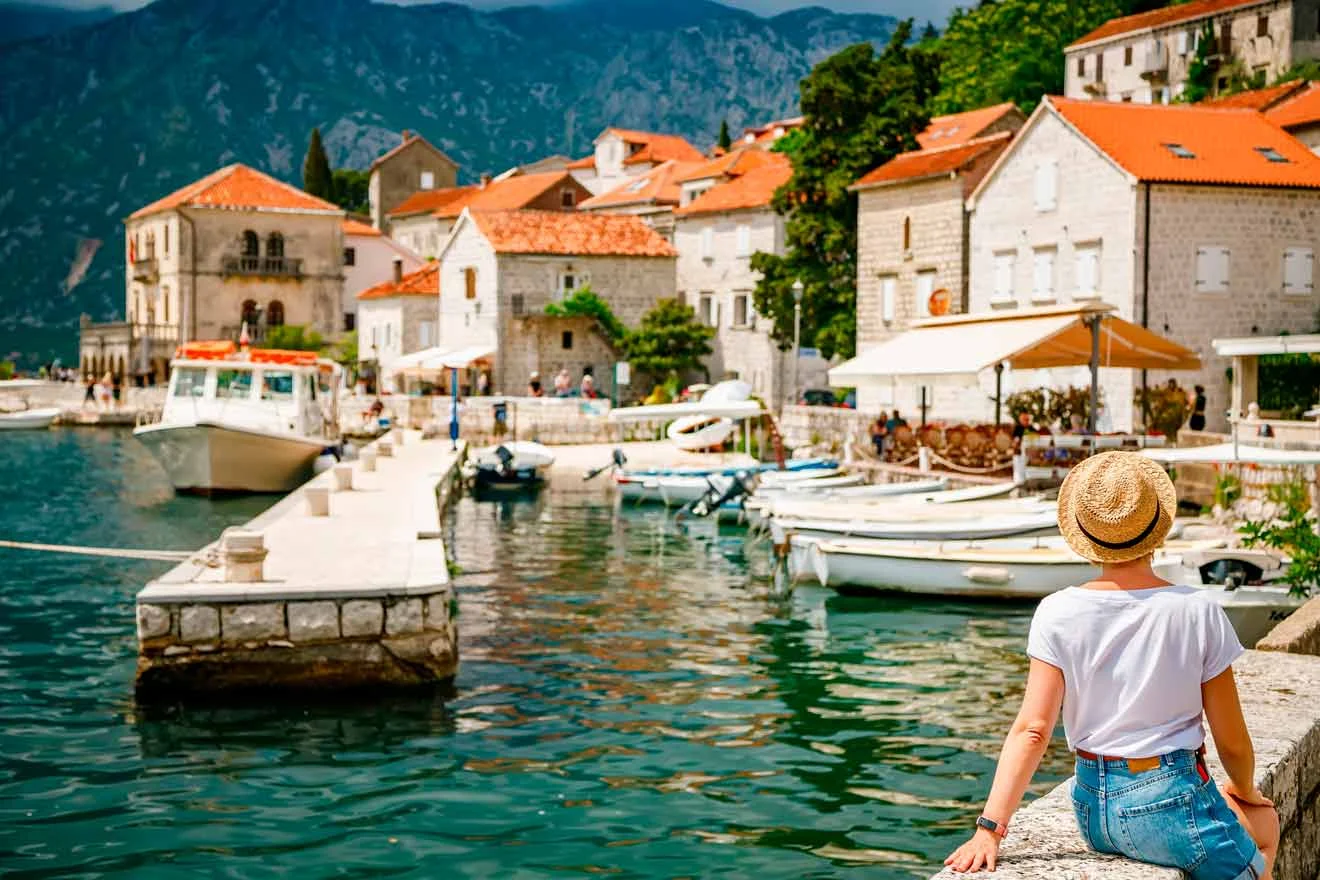 A person wearing a hat sits on a stone ledge by a marina, with boats docked and historic buildings in the background.