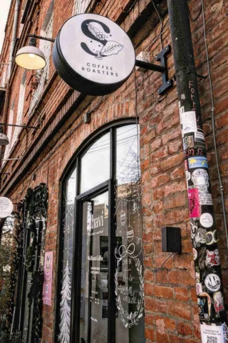 Exterior of a brick coffee shop with a round sign reading "Coffee Roasters." The entrance is glass with decorative artwork. A lamppost nearby is covered with stickers.