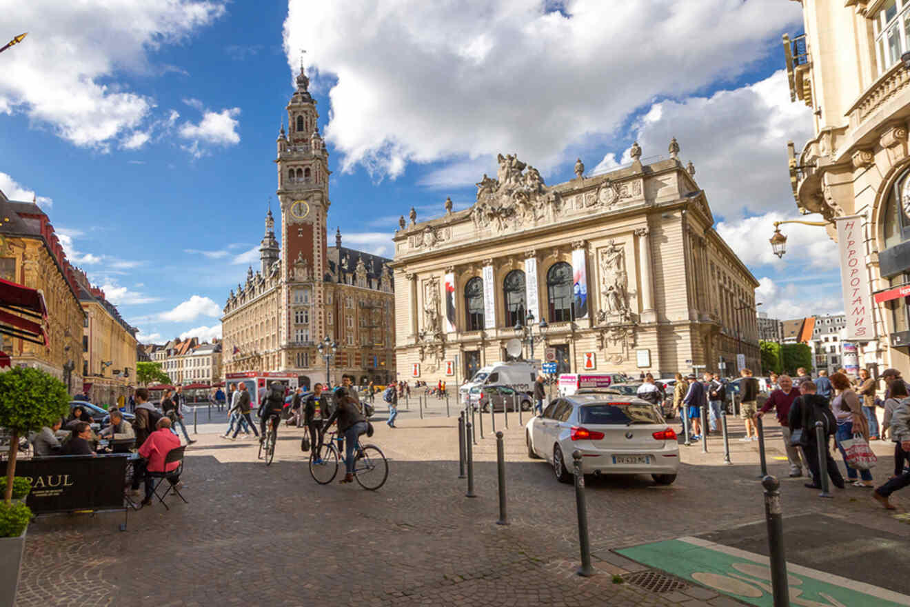 People walking and cycling in a busy plaza with historic buildings, including a clock tower and an ornate theater, under a partly cloudy sky.