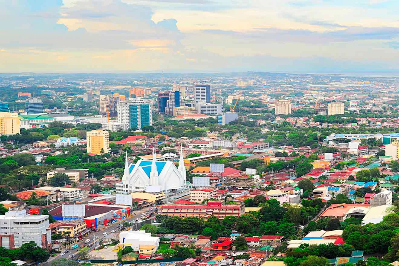 Aerial view of a cityscape with modern buildings, residential areas, and green spaces under a partly cloudy sky.